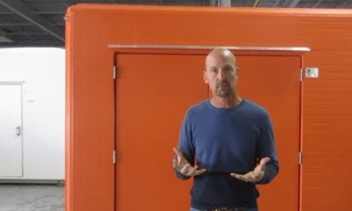 still frame from video - a man standing in front of a bright, highly visible orange shelter
