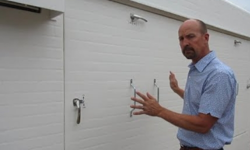 still frame from video - a man stands outside a shelter unit gesturing to where optional access panels can be placed in the exterior walls of the shelter