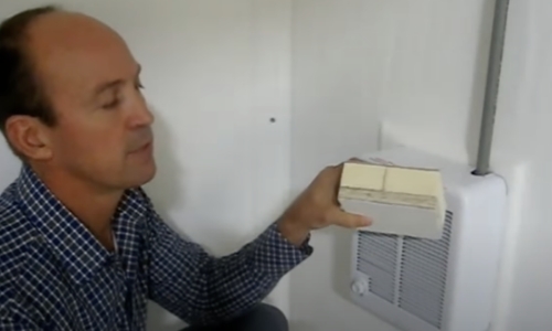 still frame from video - a man holds a cross sectioned portion of a shelter wall, showing the materials and construction of the shelter
